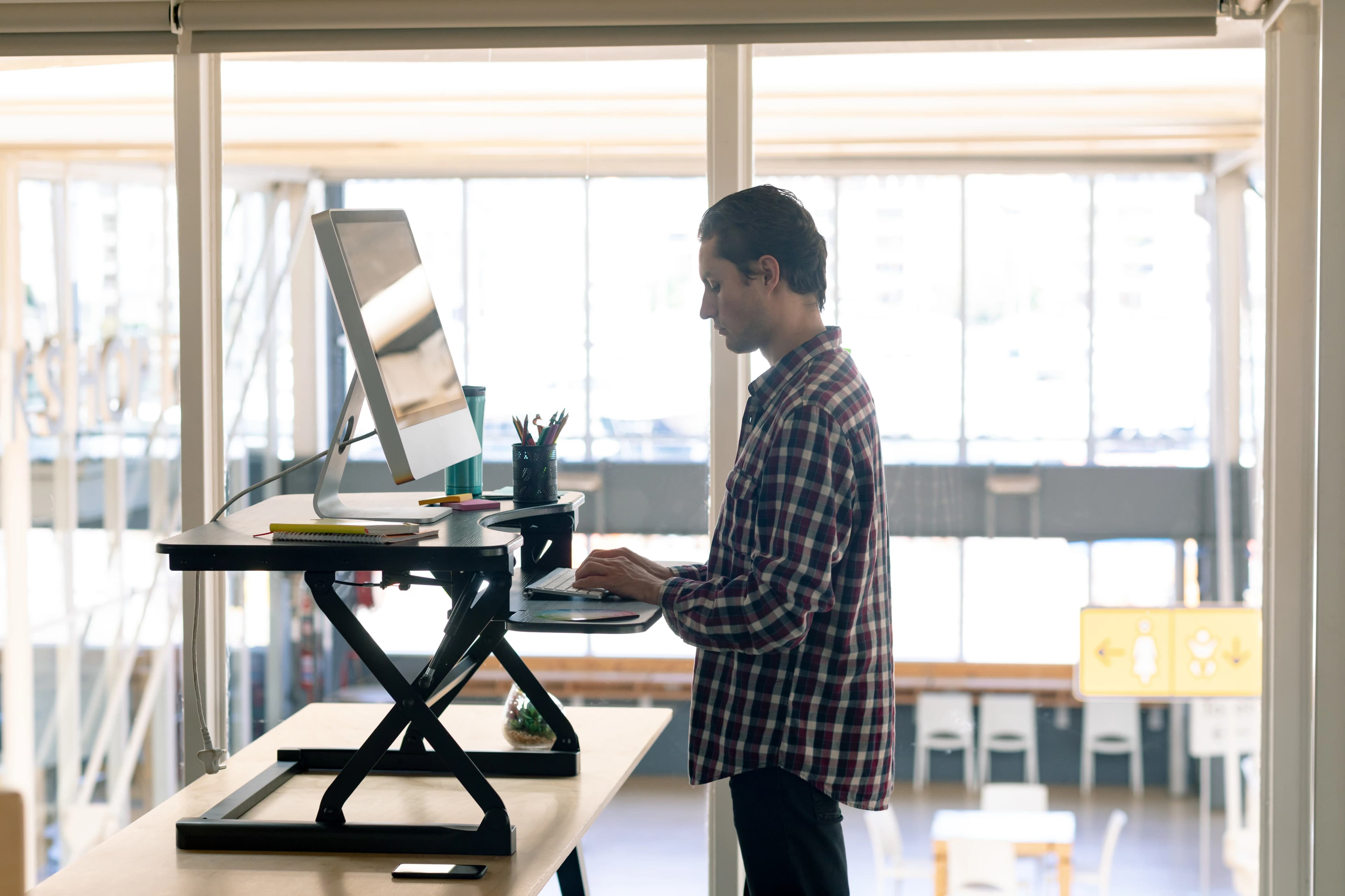 A person in a plaid shirt stands at a desk using a computer, with a bright, spacious office background. The setting is modern and productive.
