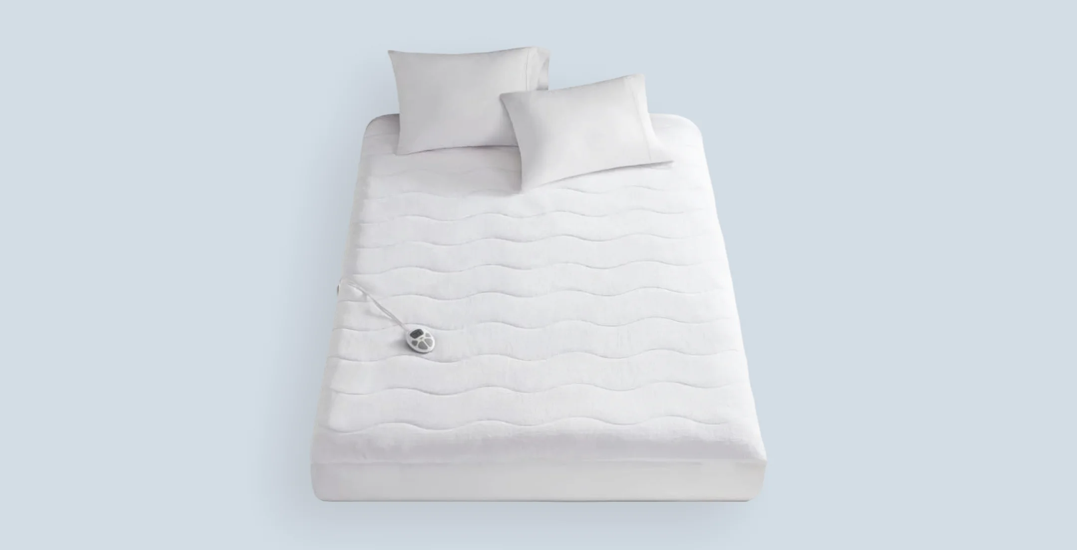 A neatly made white bed with two pillows on it. A corded remote rests on the mattress, indicating an electric blanket. The setting is minimal and clean.