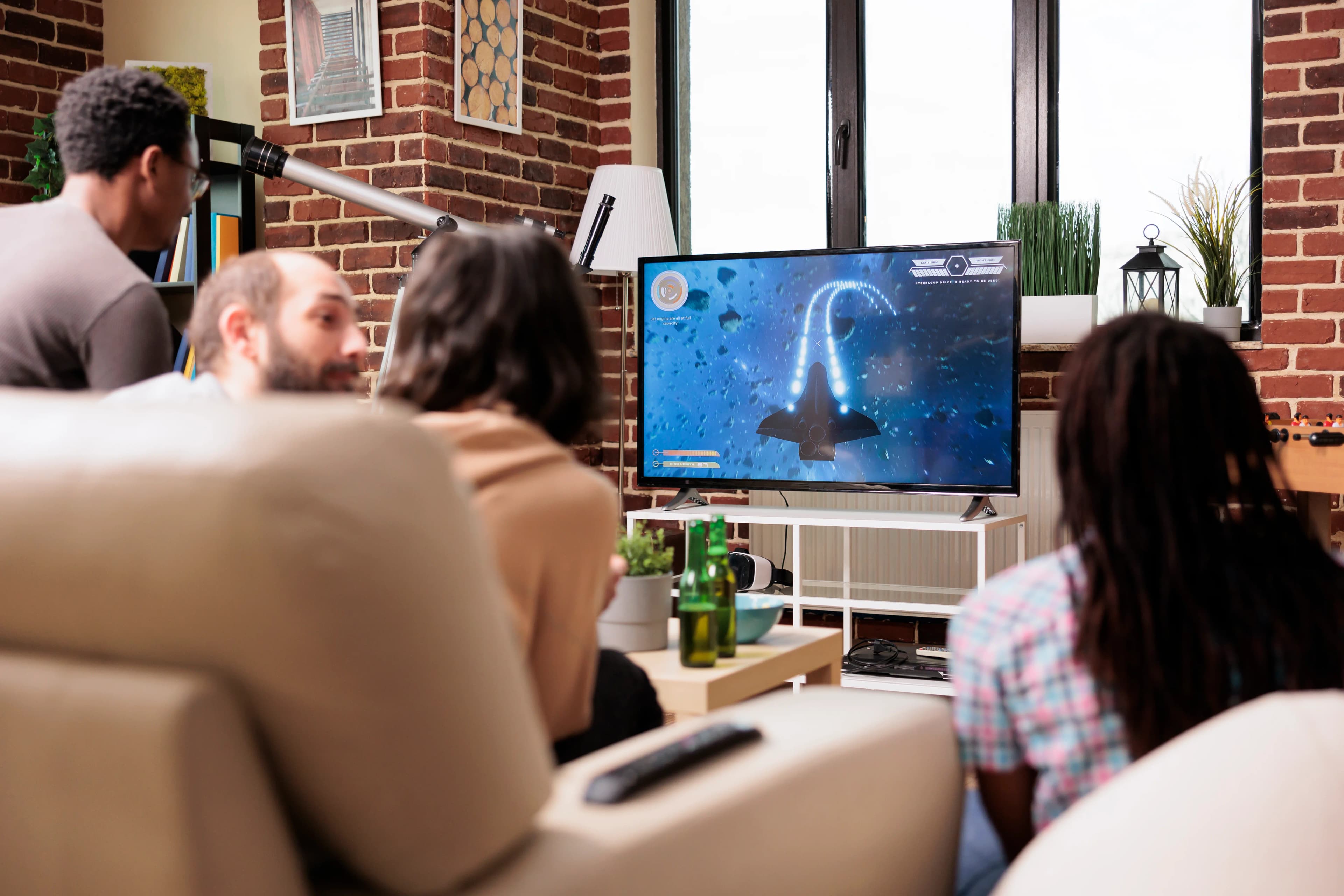 A folded blue electric blanket with a control is placed on a bed with brown, white, and patterned pillows and a gray knitted throw blanket.