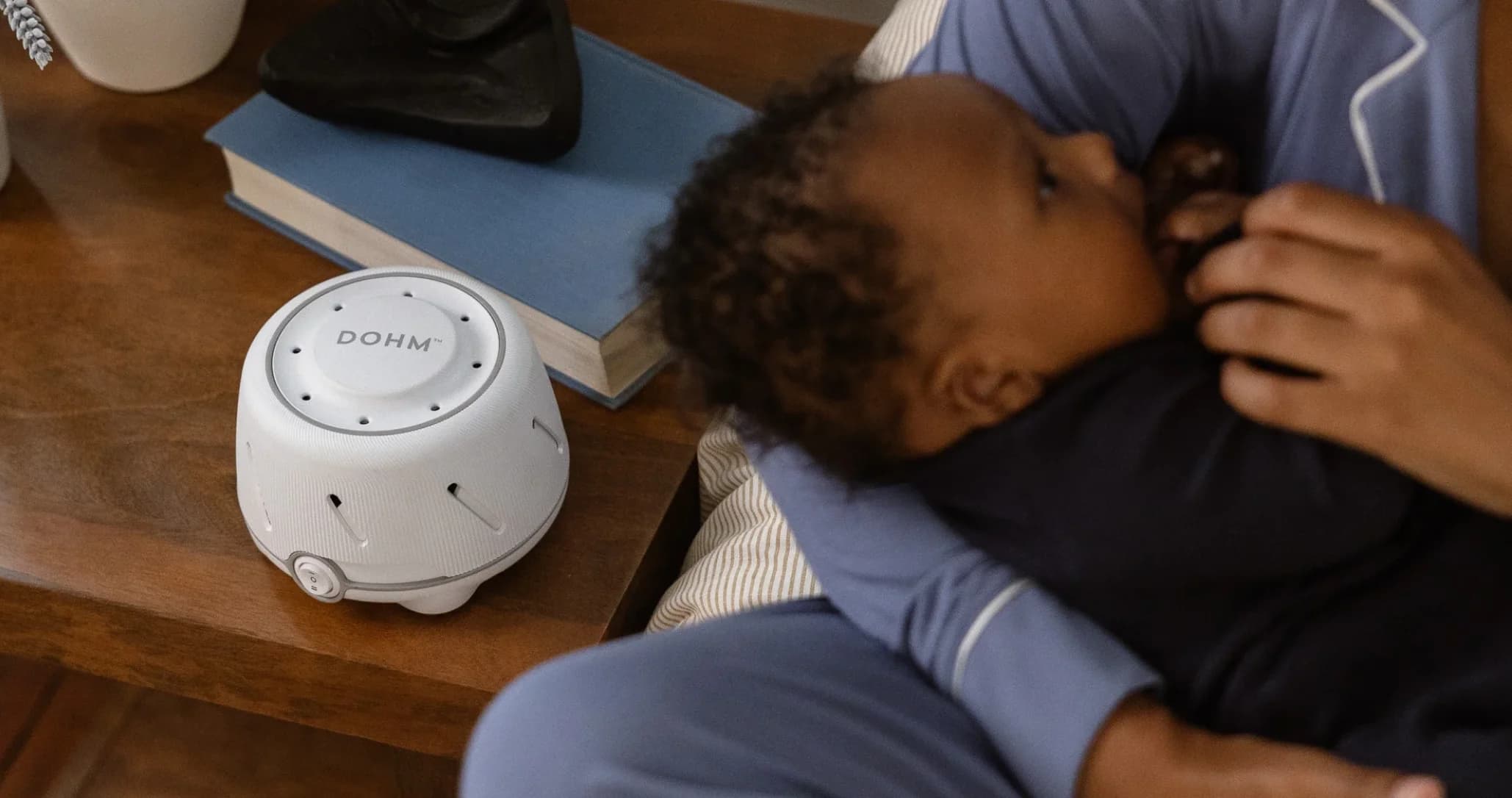 A baby is held closely by a person in blue pajamas, evoking comfort. Nearby is a white Dohm sound machine on a wooden table with a book and black pouch.